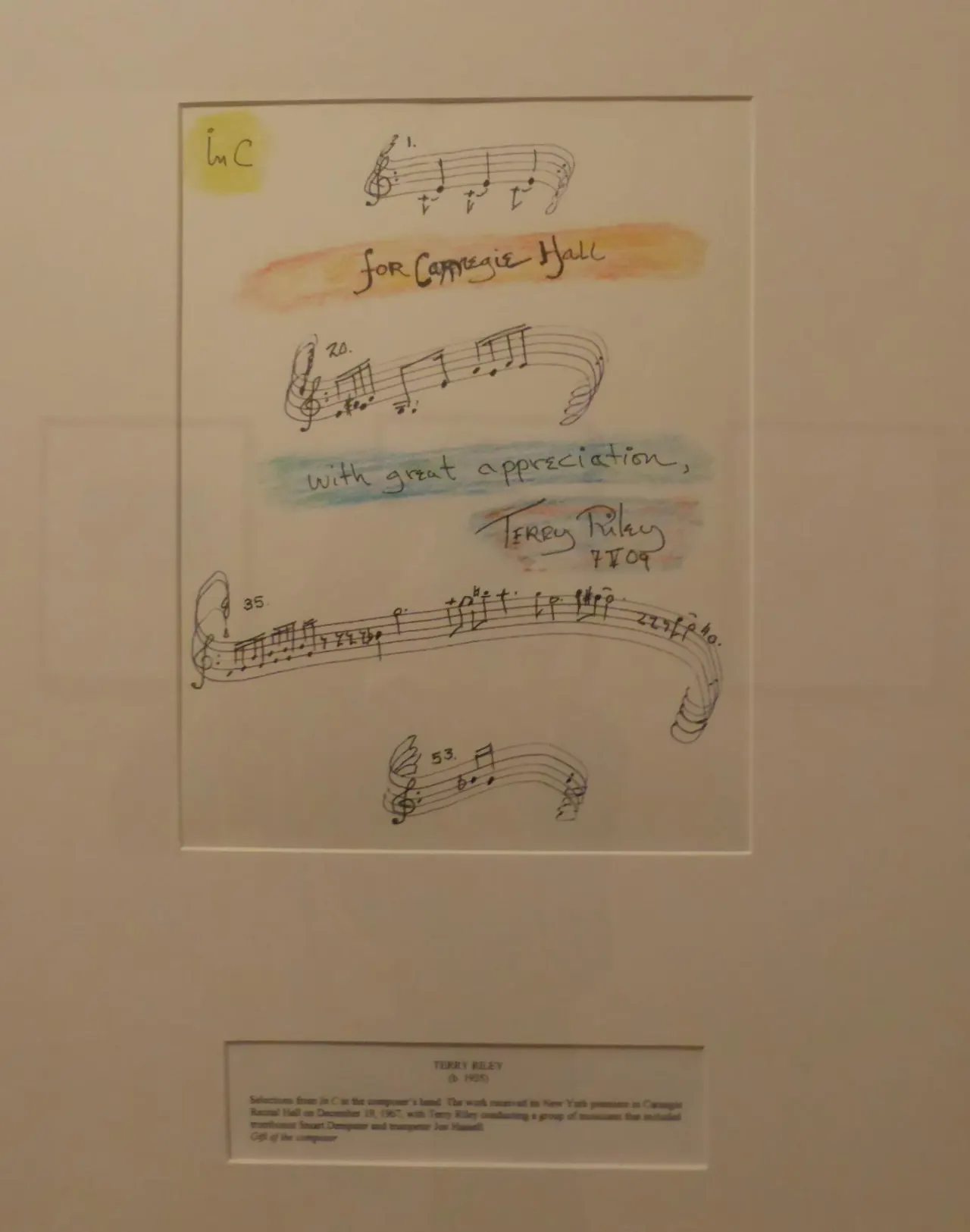 Encadr&eacute;e sur un mur du Carnegie Hall, une page de musique manuscrite de la main de Terry Riley : quelques port&eacute;es de motifs courts, rehauss&eacute;es &agrave; l'aquarelle, portant la d&eacute;dicace &laquo; for Carnegie Hall, with great appreciation &raquo; et sa signature