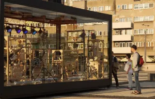 Un pavillon vitr&eacute; pos&eacute; sur une place ; &agrave; l'int&eacute;rieur, un assemblage de bois, de cordes et de poulies reli&eacute; &agrave; une batterie et &agrave; un clavier ; trois passants regardent &agrave; travers la vitre