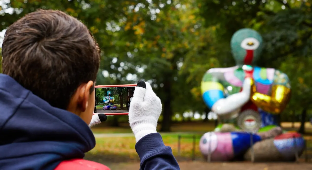 Un visiteur de dos l&egrave;ve son t&eacute;l&eacute;phone vers une sculpture color&eacute;e pos&eacute;e dans l'herbe du parc ; l'&eacute;cran affiche la silhouette &agrave; faire co&iuml;ncider avec elle