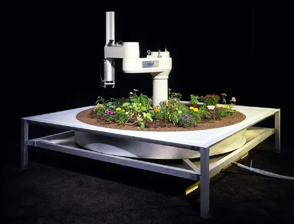 Un bras robotis&eacute; industriel mont&eacute; au centre d'un parterre de terre circulaire pos&eacute; sur une table d'acier, plant&eacute; de fleurs et de pousses, sur fond noir