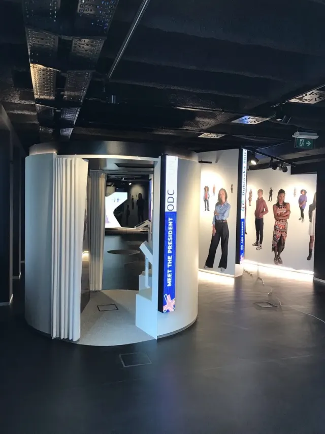 Vue d'une salle en cours d'installation, &eacute;clairage &eacute;teint. Au centre, une cabine cylindrique blanche ferm&eacute;e par un rideau &agrave; plis, ouverte sur un c&ocirc;t&eacute; ; &agrave; son flanc, un bandeau bleu vertical &laquo; MEET THE PRESIDENT &raquo; et un petit pupitre inclin&eacute;. &Agrave; droite, une paroi blanche couverte de silhouettes de personnes d&eacute;coup&eacute;es grandeur nature, &eacute;clair&eacute;e par un liser&eacute; au sol. Un c&acirc;ble tra&icirc;ne sur le sol de b&eacute;ton.