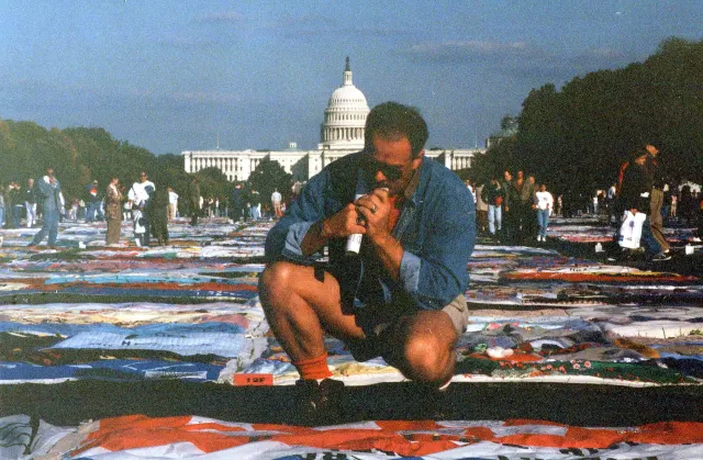 Une immense &eacute;tendue de panneaux de tissu de toutes les couleurs couvre le gazon du National Mall jusqu'au Capitole de Washington, au fond. Des visiteurs marchent dans les all&eacute;es entre les panneaux. Au premier plan, un homme en veste en jean et lunettes de soleil est accroupi au bord d'un panneau.