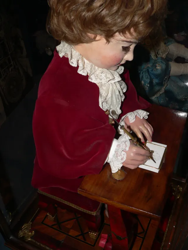 Un automate en forme de petit gar&ccedil;on aux cheveux boucl&eacute;s, veste de velours rouge et col de dentelle, assis &agrave; une table de bois. Sa main droite tient un crayon mont&eacute; sur une tige de laiton et trace un portrait sur une petite carte blanche. &Agrave; l'arri&egrave;re-plan, dans la p&eacute;nombre d'une vitrine, la robe bleue d'un autre automate.