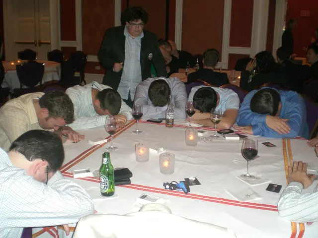 Une dizaine d'hommes en chemise assis autour d'une table napp&eacute;e de blanc, la t&ecirc;te pos&eacute;e sur les bras. Sur la table, des verres de vin, des bouteilles de bi&egrave;re, des photophores et de petites cartes retourn&eacute;es. Un jeune homme en veste sombre se tient debout derri&egrave;re eux et les regarde.