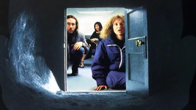 Vue depuis l'int&eacute;rieur d'un tunnel sombre aux parois de terre humide. Au bout, une petite porte grise ouverte sur un bureau &eacute;clair&eacute; au n&eacute;on. Un homme barbu &agrave; lunettes et une femme aux cheveux boucl&eacute;s, en surv&ecirc;tement bleu, sont accroupis devant l'ouverture et regardent dedans, l'air inquiet. Derri&egrave;re eux, une femme brune assise sur une chaise sourit.